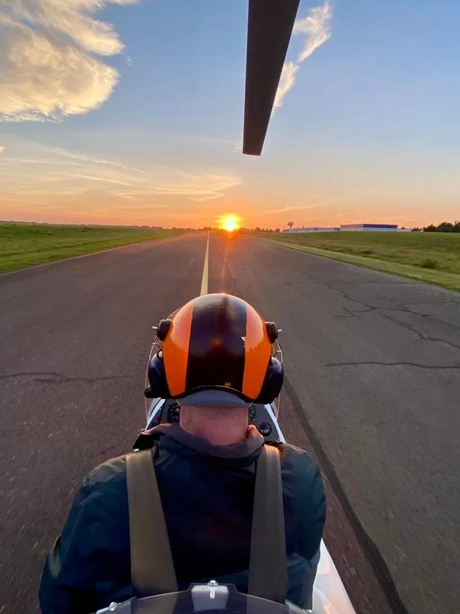 View from the back seat of a gyrocopter after landing: Christoph with an orange helmet on the taxiway, the rotor blade above, the sun just about to set